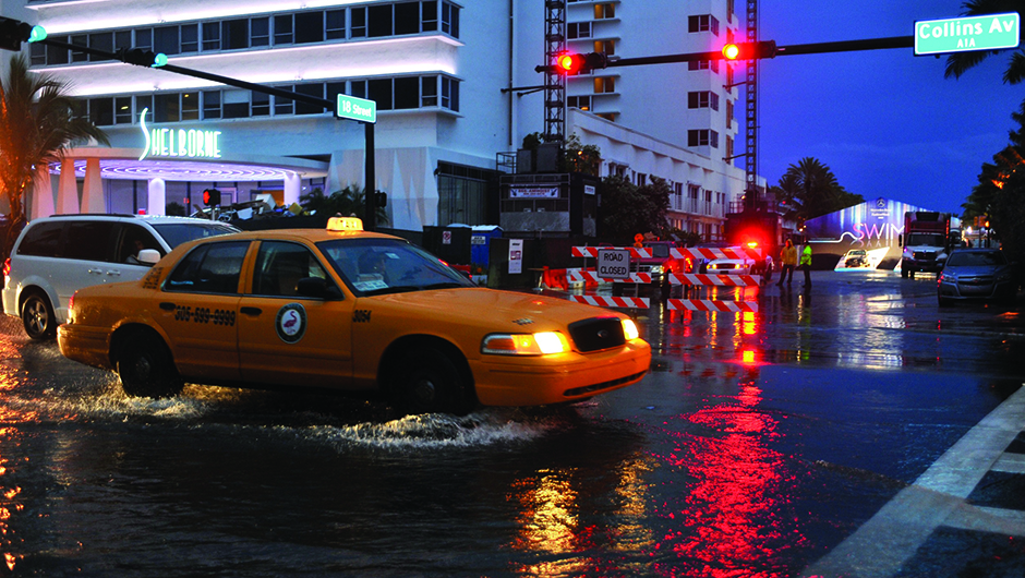 a yellow taxi ploughs through flood waters in front of the Shelbourne Hotel on Collins Avenue in Miami Beach. Headlights and traffic lights are reflected on the water's surface. A "SWIM" popup store can be seen in the background.